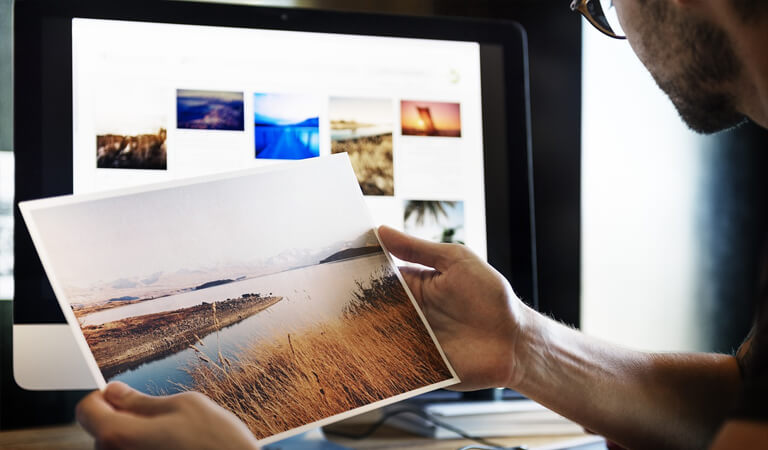 A man sitting at his desk holding a beautiful picture of a natural river and grassland while peering over to a vision board-like collection of images with similar settings on his desktop computer screen